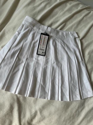 Vit plisserad kjol från FB Sister - Snygg vit plisserad kjol från FB Sister med hög midja och dragkedja i sidan. Kjolen har klassisk tennisstil och är tillverkad i ett mjukt tyg som ger en skön passform. Perfekt för en trendig och sportig look.