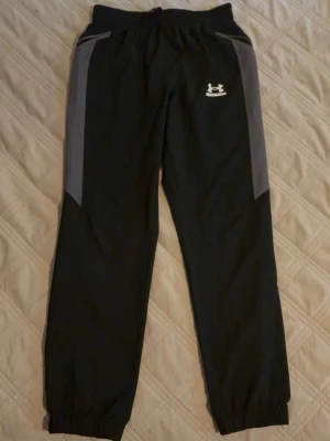 Svarta mjukisbyxor från Under Armour - Snygga svarta mjukisbyxor från Under Armour med gråa paneler längs sidorna och resår i midja och benslut. Byxorna har en avslappnad passform och är perfekta för chill eller träning. Tillverkade i ett mjukt och lätt material med logga på vänster lår.