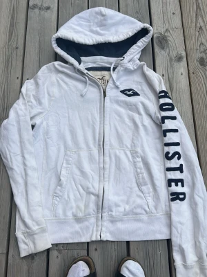 Vit hoodie från Hollister med tryck - Vit hoodie med dragkedja från Hollister. Stor logga i mörkblått på ena ärmen och Hollister-märke på bröstet. Mjuk insida, två fickor fram och justerbar huva med snören. Perfekt för en avslappnad stil.