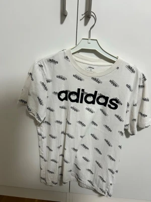 Vit Adidas t-shirt med logotypmönster storlek S - Säljer en vit t-shirt från Adidas med svart logotyptryck över hela tröjan och stor Adidas-logga på bröstet. Klassisk rund hals och korta ärmar. Perfekt för dig som gillar sportig och trendig stil.