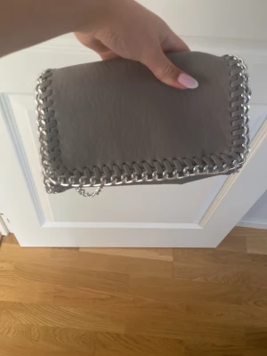 Grå handväska med kedjedetaljer - Snygg grå handväska från Tiamo med silverfärgade kedjedetaljer runt kanterna. Väskan har en rektangulär form och är gjord i ett slätt konstläder. Invändigt finns ett fack med dragkedja för småsaker. Perfekt accessoar för att lyfta din outfit. Pris kan diskuteras❤️