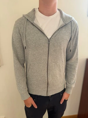 Ljusgrå zip hoodie med huva - Säljer en ljusgrå Jack & Jones hoodie i storlek XS. Den passar bra för dig som är smal eller extra smal och vill ha en lite tajtare passform. Den är i väldigt bra skick och är riktigt skön att ha på sig.  Perfekt nu till skolstarten och enkel att matcha med typ jeans eller mjukisbyxor. Funkar lika bra i skolan som när man bara chillar hemma eller är ute med kompisar. Har dragkedja framtill, två fickor och justerbara snören i huvan.  En enkel och stilren hoodie som passar till det mesta.