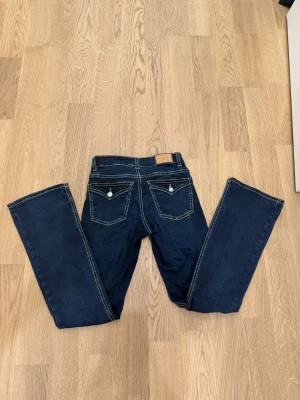 Mörkblå bootcut jeans med kontrastsömmar - Snygga mörkblå jeans med bootcut passform och vita kontrastsömmar. Bakfickorna har lock och silverfärgade knappar som detalj. Klassisk femficksmodell i mjukt denimtyg, perfekta till en avslappnad look.