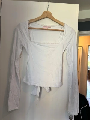 Vit långärmad topp med knyt - Snygg vit långärmad topp med fyrkantig ringning och knyt i midjan. Toppen har en feminin touch med spetsdetalj längs urringningen och är figurnära i modellen. Perfekt att styla med jeans eller kjol.