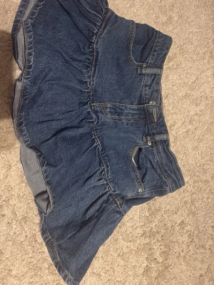 Blå jeanskjol från Gina Tricot  - Snygg blå jeanskjol från Perfect Jeans i kort modell. Kjolen har volang nertill, klassiska jeansfickor och synliga sömmar. Perfekt för dig som gillar en avslappnad och trendig stil.