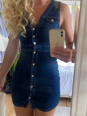 Ärmlös jeansklänning med knappar - Snygg ärmlös jeansklänning i blå denim med v-ringning och knappar hela vägen framtill. Två bröstfickor med lock och kontrastsömmar ger en cool retrovibe. Perfekt till sommaren och lätt att styla med t-shirt eller tröja under.