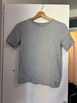 Grå Ralph Lauren T-shirt - En enkel grå t-shirt med rund hals och korta ärmar. Tillverkad i mjuk bomull, perfekt för en avslappnad stil. T-shirten har en diskret broderad detalj nertill på framsidan.