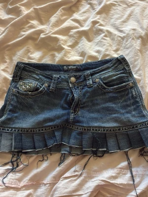 Låg midjad Jeanskjol med paljettdetaljer  - Cool jeanskjol i blå denim med plisserad nederkant och slitna fransar. Fickor fram och bak, dekorerade med paljetter och broderier. Perfekt för en edgy och trendig look. kjolen ser exakt ut så som den såg ut när jag köpte den, med trådarna och hur loggan ser ut som är beige färgad, Den här kjolen köpte jag från A2 Vintage i mall of Scandinavia. 2025 sommaren, Pris går såklart att diskutera 