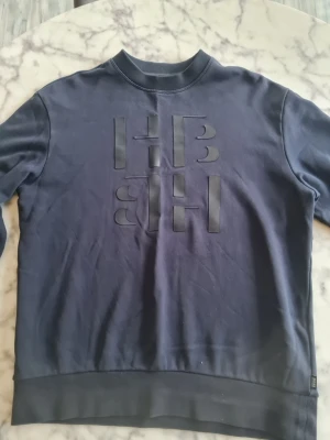 Mörkblå Hugo Boss sweatshirt med tryck - Mörkblå Hugo boss sweatshirt med rund hals och stora, upphöjda bokstäver 'HB' framtill. Tröjan har långa ärmar och breda muddar vid ärmslut och nederkant. Perfekt för en avslappnad och stilren look.