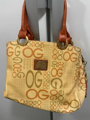 Beige och orange handväska Goldfish - Cool handväska från Goldfish i beige tyg med orange och bruna bokstäver och logga. Väskan har bruna handtag i konstläder och stora guldfärgade ringar. Perfekt för dig som gillar retrostil och vill sticka ut lite extra.