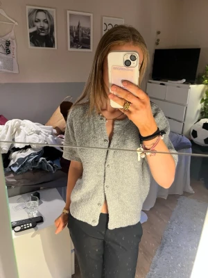 Grå kortärmad kofta från Gina Tricot - Mysig grå kofta från Gina Tricot med korta ärmar och rund halsringning. Koftan har en fluffig, texturerad yta och knäpps framtill med små knappar. Perfekt att slänga över en topp för en chill look.