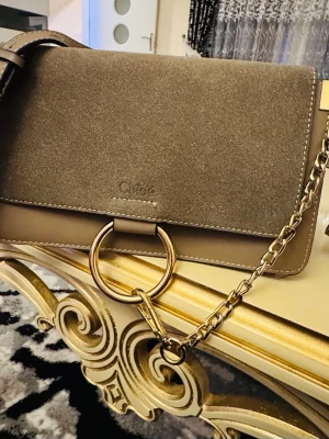 Beige handväska från Chloé - Snygg beige handväska från Chloé i mocka och läder. Väskan har guldfärgade metalldetaljer, en kedjerem och en stor ring framtill. Perfekt accessoar för dig som gillar lyxiga detaljer och stilren design.