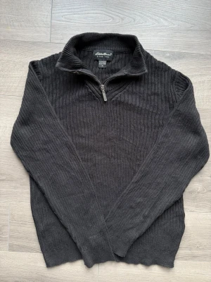 Svart stickad tröja från Eddie Bauer - Säljer en svart stickad tröja från Eddie Bauer i 100% pima bomull. Tröjan har halv dragkedja, ribbad struktur och hög krage. Perfekt för dig som gillar klassisk och enkel stil med en modern twist.