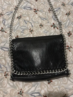 Svart axelväska med kedjedetaljer Tiamo - Snygg svart axelväska från Tiamo i konstläder med grova silverfärgade kedjedetaljer runt kanterna och som axelrem. Väskan har lock och innerfack med dragkedja. Perfekt för dig som gillar en edgy och trendig look.