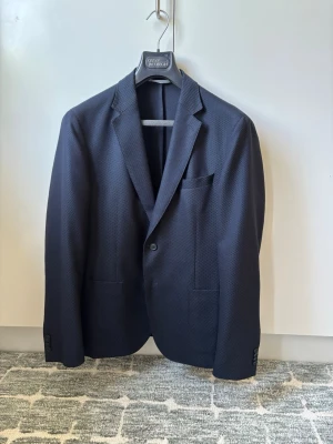 Mörkblå strukturerad kavaj från Hugo Boss - Snygg mörkblå kavaj med diskret strukturmönster och klassisk passform. Kavajen har två framfickor, bröstficka och två knappar vid ärmslut. Perfekt för en stilren look och enkel att matcha med skjorta eller t-shirt.