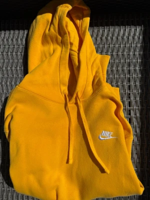 Gul hoodie från Nike med huva - Snygg gul hoodie från Nike med klassisk huva och dragsnören. Broderad vit Nike-logga på bröstet ger en clean look. Tillverkad i mjukt material som känns skönt mot huden. Perfekt för dig som vill sticka ut med färg i vardagen.