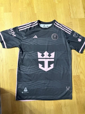 Inter Miami Messi fotbollströja - Snygg Inter Miami matchtröja från Adidas i mörkgrått med rosa detaljer. Tröjan har Messi och nummer 10 på ryggen, klubbmärke och sponsorloggor på bröst och ärmar. Tillverkad i lätt och ventilerande funktionsmaterial, perfekt för fotboll eller träning. T-shirten är ifrån 2025. Storleken är L man funkar för både L och M