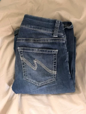 Mörkblå bootcut jeans - Säljer ett par mörkblå bootcut jeans med snygg vit brodyr på bakfickorna. 🤍. Perfekta för en avslappnad och trendig look. Jeansen har tyvvär ett litet hål/ slitning längst ner som knappt syns och som många uppfattar som snyggt, kontakta för fler bilder❣️