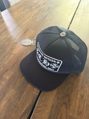 Snygg keps - Säljer en svart truckerkeps från Chrome Hearts med vit broderad patch framtill och metallknapp med logga på toppen. Kepsen har mesh på sidorna och justerbar snapback. Perfekt för dig som gillar streetwear och vill sticka ut.