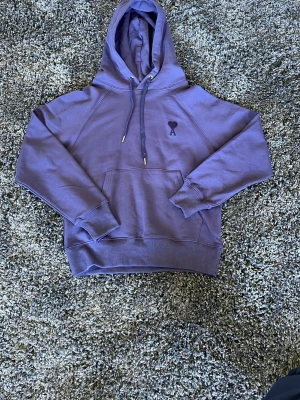 Mörkblå hoodie från Ami Paris - Snygg mörkblå hoodie från Ami Paris med klassisk huva och dragsnören. På bröstet finns Ami:s ikoniska hjärta med A-brodyr. Hoodien har en stor magficka och ribbade muddar vid ärmar och nederkant. Tillverkad i mjuk bomull för maximal komfort. Den ser lite lila ut på bilderna men är mörkblå. 