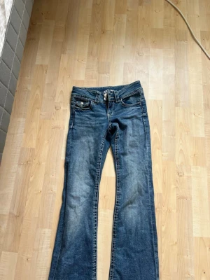 Blå bootcut jeans med slitningar - Ett par klassiska blå bootcut jeans med lätt slitna detaljer och fem fickor. Jeansen har normal midja, dragkedja och knapp framtill samt kontrastsömmar. Perfekta för en avslappnad och trendig look.