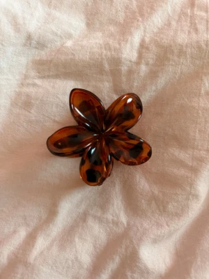 Blommig hårklämma  - Snygg hårklämma i form av en blomma  i bruna och svarta toner. Tillverkad i plast med glansig finish och stark fjäder. Perfekt för att snabbt sätta upp håret med en trendig touch.