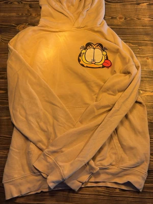 Beige hoodie med Garfield-tryck - Säljer en beige hoodie med Garfield-tryck fram och bak. På framsidan finns ett litet Garfield-ansikte och på ryggen syns Garfield på skateboard med stor röd text. Hoodien har huva och magficka, perfekt för dig som gillar streetwear och tecknade serier.