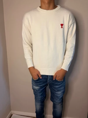 ami tröja  - Säljer en stilren vit stickad tröja med rund hals och ribbade muddar. På bröstet finns en liten röd broderad detalj i form av ett hjärta. Perfekt för dig som gillar minimalistisk design och mjuka material.