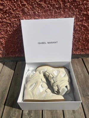 Isabel Marant vita höga sneakers - Säljer ett par vita höga sneakers från Isabel Marant med chunky sula och flera kardborreband. Skorna har en cool design i mocka och textil med detaljer i olika nyanser av vitt och beige. Perfekta för dig som vill sticka ut med din stil.