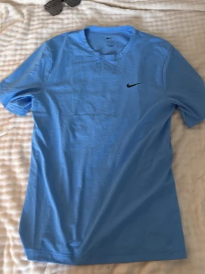 Blå Nike Dri-FIT t-shirt - Snygg ljusblå t-shirt från Nike med klassisk logga på bröstet. Tillverkad i Dri-FIT-material som andas och passar perfekt till träning. Kortärmad och med rund hals för en clean och sportig look.