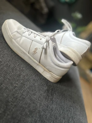 Vita låga sneakers från Polo Ralph Lauren - Klassiska vita sneakers i läder från Polo Ralph Lauren. De är använda några gånger som man kan se. Lite skitiga på sulan men väldigt fina skor som passar till det mesta! Säljer dem för lågt pris eftersom de finns lite skavanker på dem.