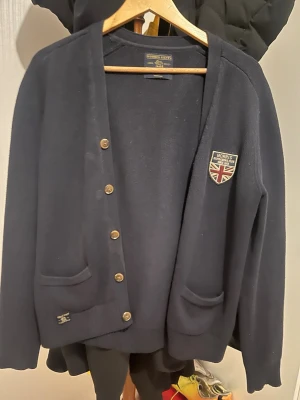 Mörkblå cardigan från Morris - Stilren mörkblå cardigan från Morris med knappar framtill och två fickor. Dekorerad med Morris-logga och brittisk flagga på bröstet. Perfekt för en klassisk och avslappnad look.
