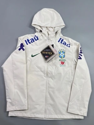Vit Brasil Gore-Tex vindjacka - Snygg vit vindjacka med huva och dragkedja framtill. Jackan har Brasil-logga, Nike-logga och flera sponsortryck i blått och grönt. Tillverkad i slitstarkt Gore-Tex-material som står emot vind och regn. Perfekt för dig som gillar sportig stil och vill sticka ut.