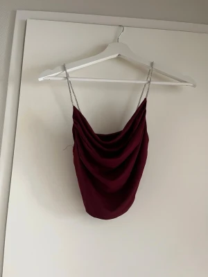 Vinröd draperad linnetopp med kedjeband - Trendig vinröd linnetopp med draperad front och tunna kedjeband som axelband. Toppen har en croppad passform och är perfekt för att skapa en snygg och stilren look. Materialet känns mjukt och följsamt mot huden.