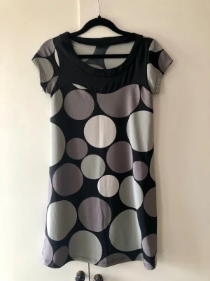 Gina Tricot Prickig Klänning / Tunika Retrostil – Stl XS - Snygg kortare klänning eller tunika från Gina Tricot i härlig retrostil med stora prickar i grå och lila toner. Modell med kort ärm och svart parti vid halsringningen. Stretchigt och skönt material. Storlek: XS Mått: Axelbredd ca 34 cm, längd ca 78 cm Material: Syntetblandning (tvättlapp saknas) Skick: Mycket gott skick