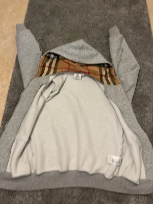 Grå hoodie med mönstrad huva - Säljer en grå zip-hoodie med klassiskt rutmönstrat innerfoder i huvan. Hoodien har dragkedja framtill, två fickor och justerbara snören i huvan. Perfekt för dig som gillar stilrena plagg med en twist. Materialet är mjukt och bekvämt, troligen bomullsblandning.