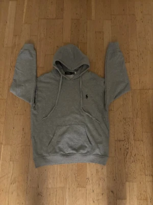 Grå hoodie från Polo Ralph Lauren - Snygg grå hoodie från Polo Ralph Lauren med klassisk huva och dragsnören. Tröjan har en stor magficka och den ikoniska svarta Polo-loggan broderad på bröstet. Perfekt för en chill och avslappnad stil. Tillverkad i mjukt bomullsmaterial. OBS! Osäker på äktighet så ha det i åtanke!