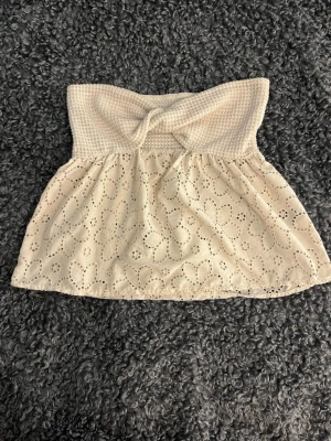 Beige axelbandslös topp med spets - Söt beige axelbandslös topp med broderade blommor och spetsdetaljer. Överdelen är smockad och har en liten rosett framtill. Perfekt för varma dagar och sommarens alla tillfällen.
