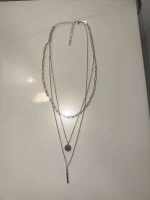 Silverfärgat lager-på-lager halsband - Snyggt silverfärgat halsband med tre lager. Det översta lagret har större länkar, det mellersta har en rund, hamrad berlock och det nedersta lagret har ett avlångt stavhänge. Är inte i äkta silver kommer från H&M.