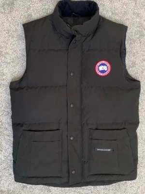 Svart dunväst från Canada Goose - Snygg svart dunväst från Canada Goose med hög krage och två stora fickor framtill. Västen har ett klassiskt Canada Goose-märke på bröstet och stängs med tryckknappar. Perfekt för lager-på-lager och håller dig varm under kyliga dagar.