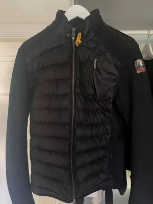 Svart parajumper cardigan - Snygg svart dunväst från Parajumpers med quiltad front och rygg, samt stickade ärmar och axlar. Dragkedja framtill, bröstficka med dragkedja och ikonisk gul detalj vid halsen. Märkespatch på ärmen. Perfekt för lager-på-lager och streetwear.