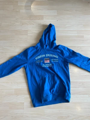 Blå hoodie från Vinson Original - Snygg blå hoodie från Vinson Original med vit text och tryck framtill. Klassisk känguruficka och huva. Skön och avslappnad passform, perfekt till jeans eller joggers. Material:bomull, polyester 