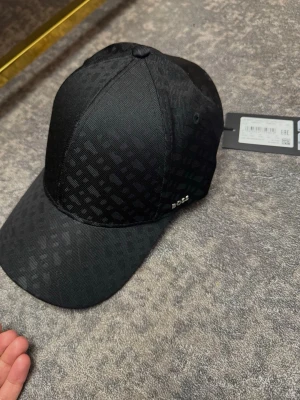 Svart keps från BOSS med mönster - Snygg svart keps från BOSS med diskret rutmönster över hela kepsen. Klassisk böjd skärm och justerbar rem bak. Tillverkad i ett slitstarkt tyg som känns både stilrent och modernt. Perfekt accessoar för en trendig look.