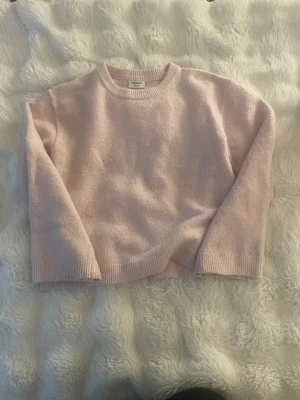 Ljusrosa stickad tröja - Mysig ljusrosa stickad tröja med rund halsringning och långa ärmar. Tröjan har ribbade muddar vid ärmslut och nederkant, vilket ger en klassisk och enkel look. Perfekt för att hålla värmen under kyliga dagar.