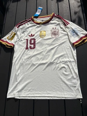 Spanien matchtröja Lamine Yamal #19 - Snygg vit Spanien matchtröja från Adidas med vinröda och gula detaljer. Tröjan har nummer 19 och Lamine Yamal på ryggen, samt officiella märken och loggor på bröstet och ärmarna. Tillverkad i lätt och ventilerande Climacool-material.