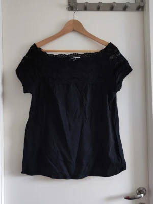 Svart topp med spetsdetalj - Snygg svart topp med bred spets upptill och korta ärmar. Toppen har en feminin offshoulder-look tack vare spetsen och är perfekt för dig som gillar stilrena plagg med en twist. Materialet känns lätt och luftigt.