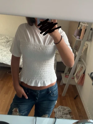 Vit smockad topp med volangkant - Säljer en söt vit topp med smockad midja och volangkant nedtill. Toppen har korta ärmar och rund halsringning. Perfekt för dig som gillar en feminin och trendig stil.