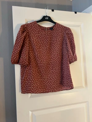 Brunprickig blus med puffärm - Söt brun blus med vita prickar och puffiga trekvartsärmar. Blusen har rund halsringning och knäppning bak i nacken. Tillverkad i ett lätt och mjukt tyg som ger en avslappnad men trendig look.