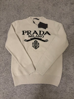 Vit stickad tröja från Prada - Snygg vit stickad tröja från Prada med svart logotyp och texten 'PRADA MILANO' framtill. Tröjan har rund halsringning, ribbade muddar och nederkant. Perfekt för dig som vill ha en exklusiv och trendig look. 