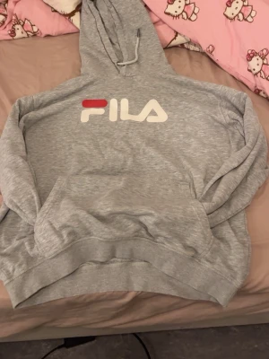 Grå hoodie från FILA med logga - Snygg grå hoodie från FILA med stor vit och röd logga på bröstet. Klassisk känguruficka framtill och justerbar huva med dragsko. Tillverkad i mjukt sweatshirtmaterial som är perfekt för chill dagar.